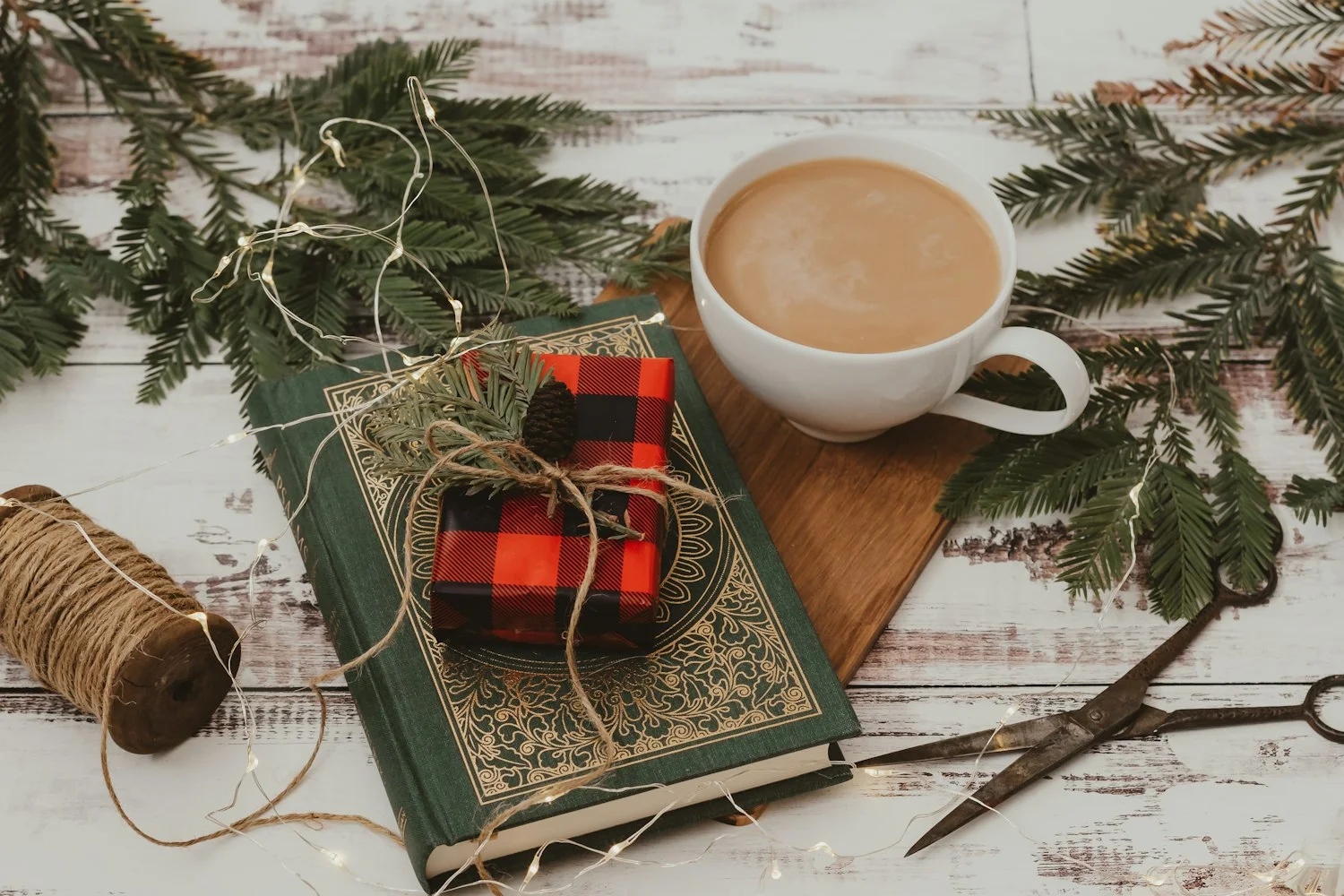 A plaid-wrapped gift tied with twine sits on a green and gold hardcover book beside a white cup on a wooden board, with evergreen sprigs and fairy lights on a whitewashed table