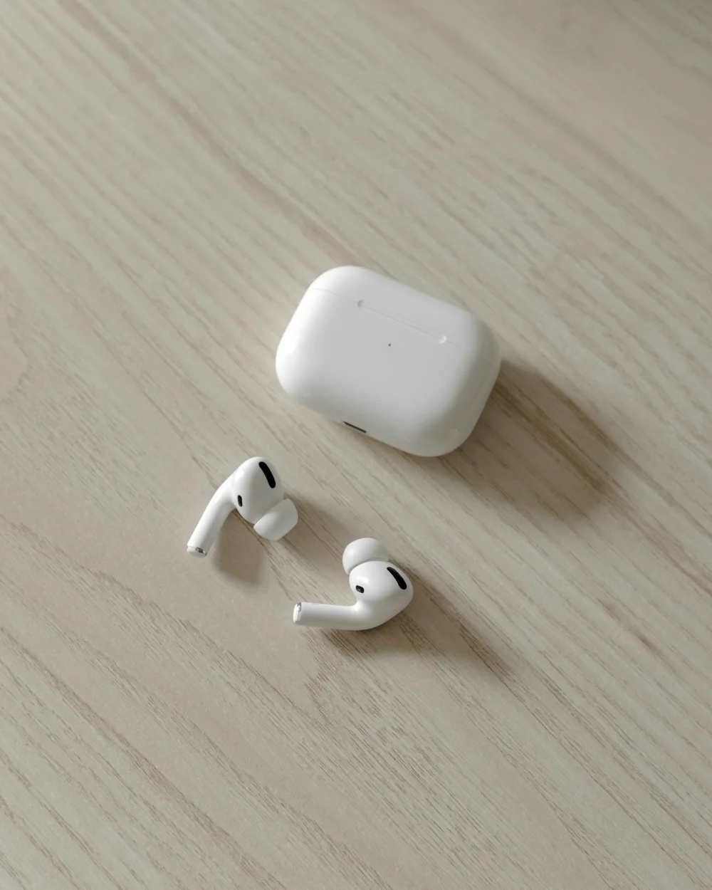 A pair of white in-ear AirPods Pro with silicone tips beside their closed charging case on a pale wood table