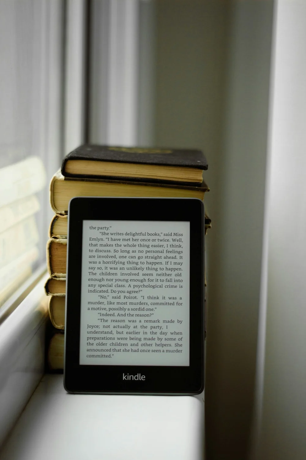 A Kindle Paperwhite showing a page of a Poirot mystery, propped against a stack of worn hardcovers on a white windowsill