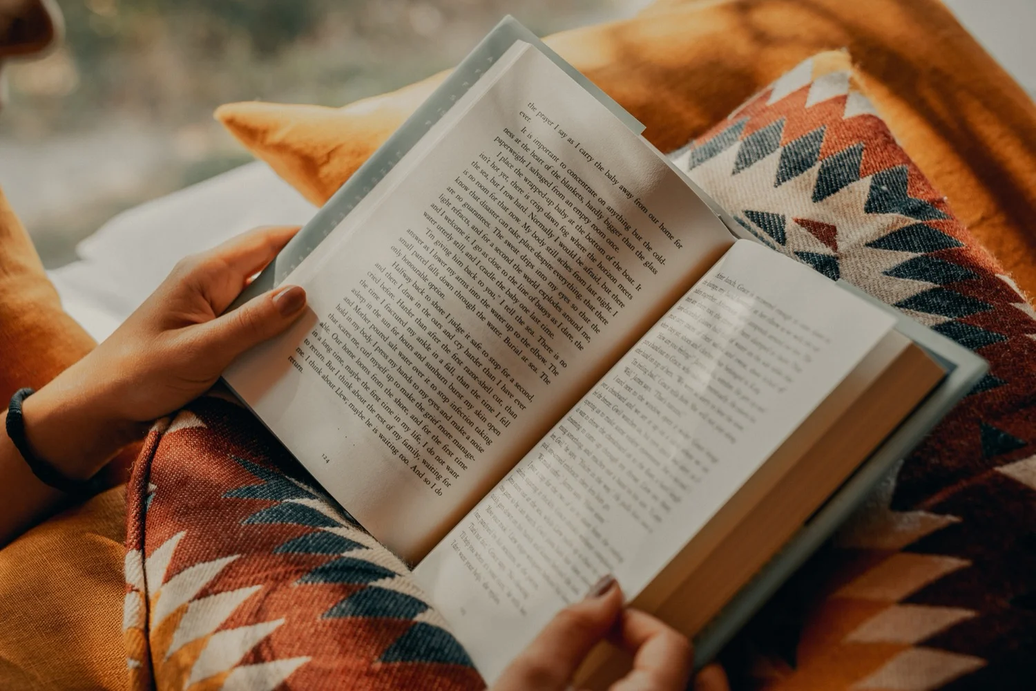 An open hardcover book in a reader’s hands on a cozy patterned blanket