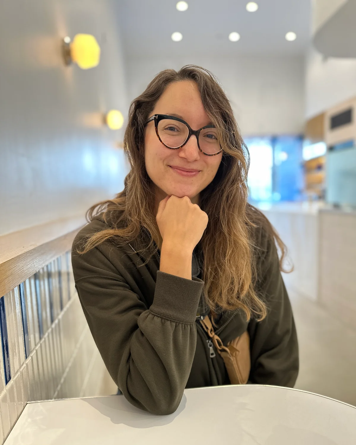 Amy Suto smiling with her chin on her hand at a white cafe table in Los Angeles, wearing an olive sweatshirt and tortoiseshell cat-eye glasses