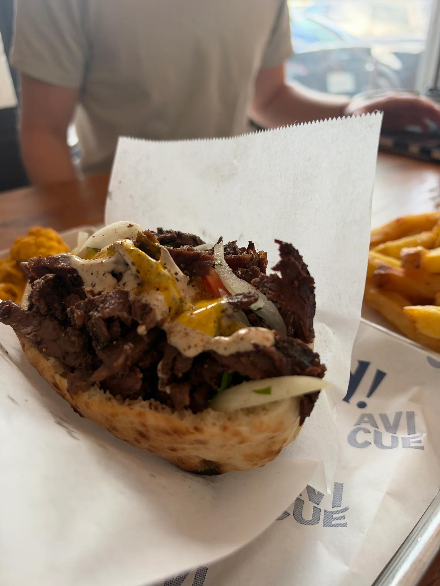 The OG Wagyu beef shawarma half pita on Avi Cue branded tray paper in Studio City, with fries on the side and Kyle out of focus behind it