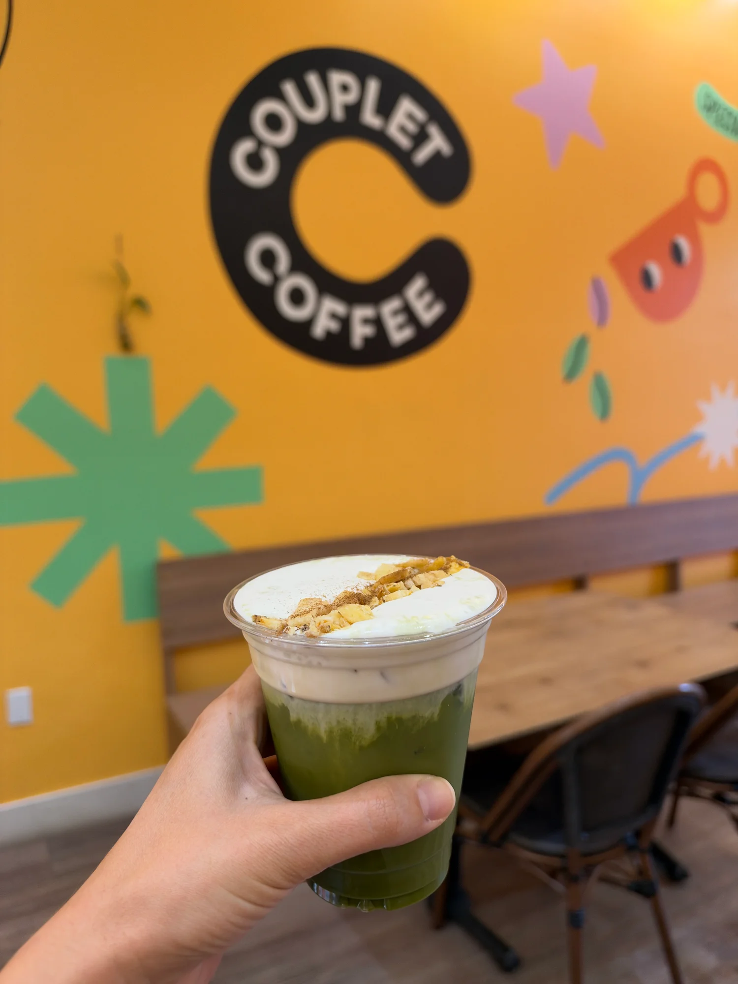 An iced banana cream matcha topped with cream and crushed banana chips, held up in front of the Couplet Coffee logo on a mustard-yellow wall at the Hollywood cafe in Los Angeles