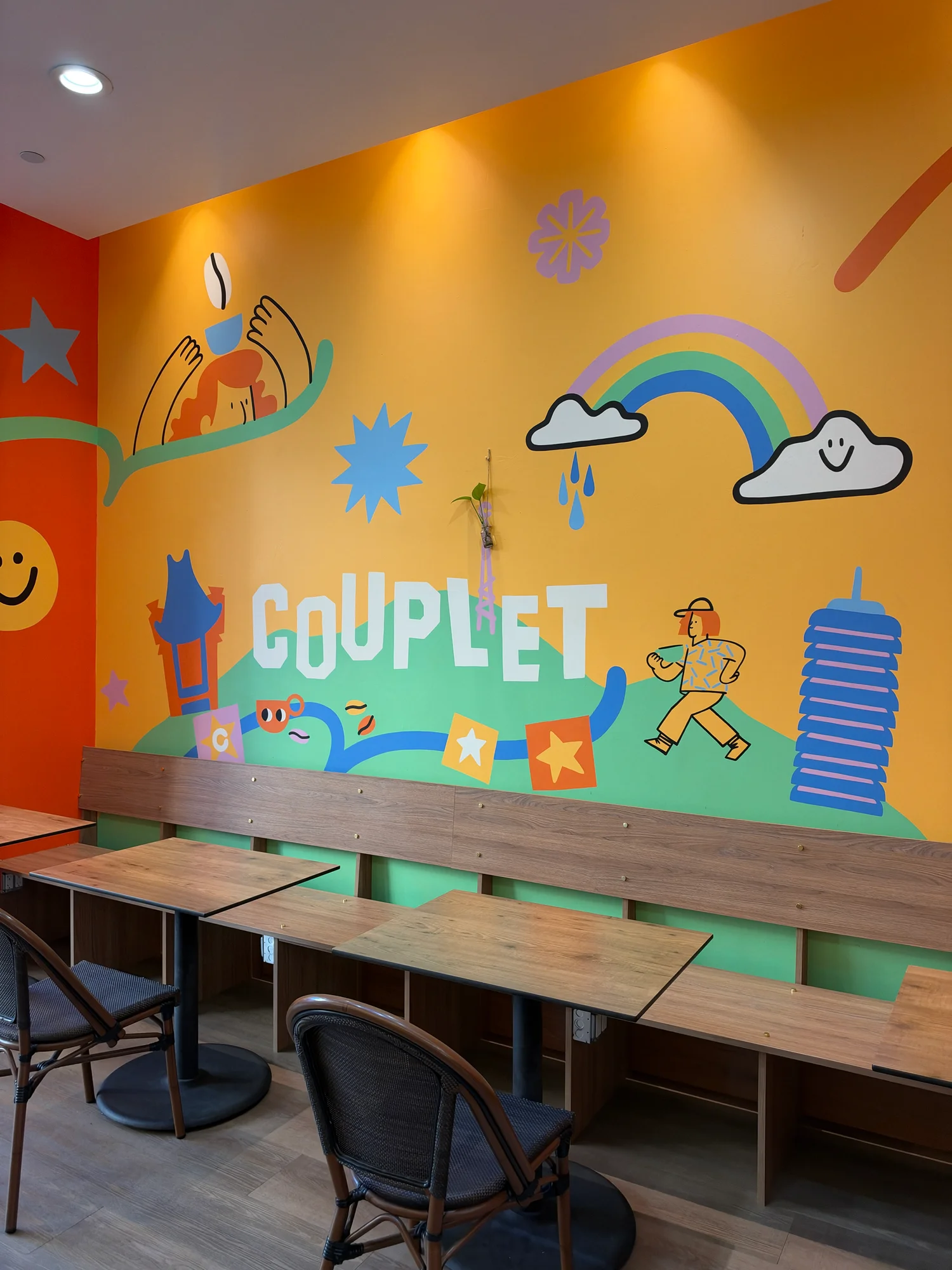 The mustard-yellow mural wall inside Couplet Coffee in Hollywood, painted with a rainbow, a smiling cloud, a pagoda-roofed tower and the word COUPLET above a long wooden bench and small tables