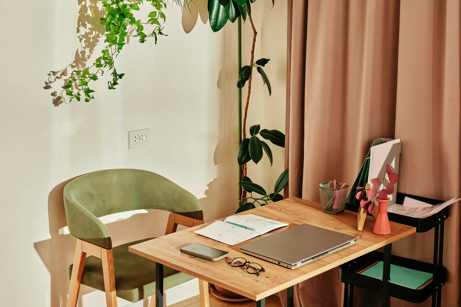 A laptop and notes on a light wooden desk with a green chair and plants