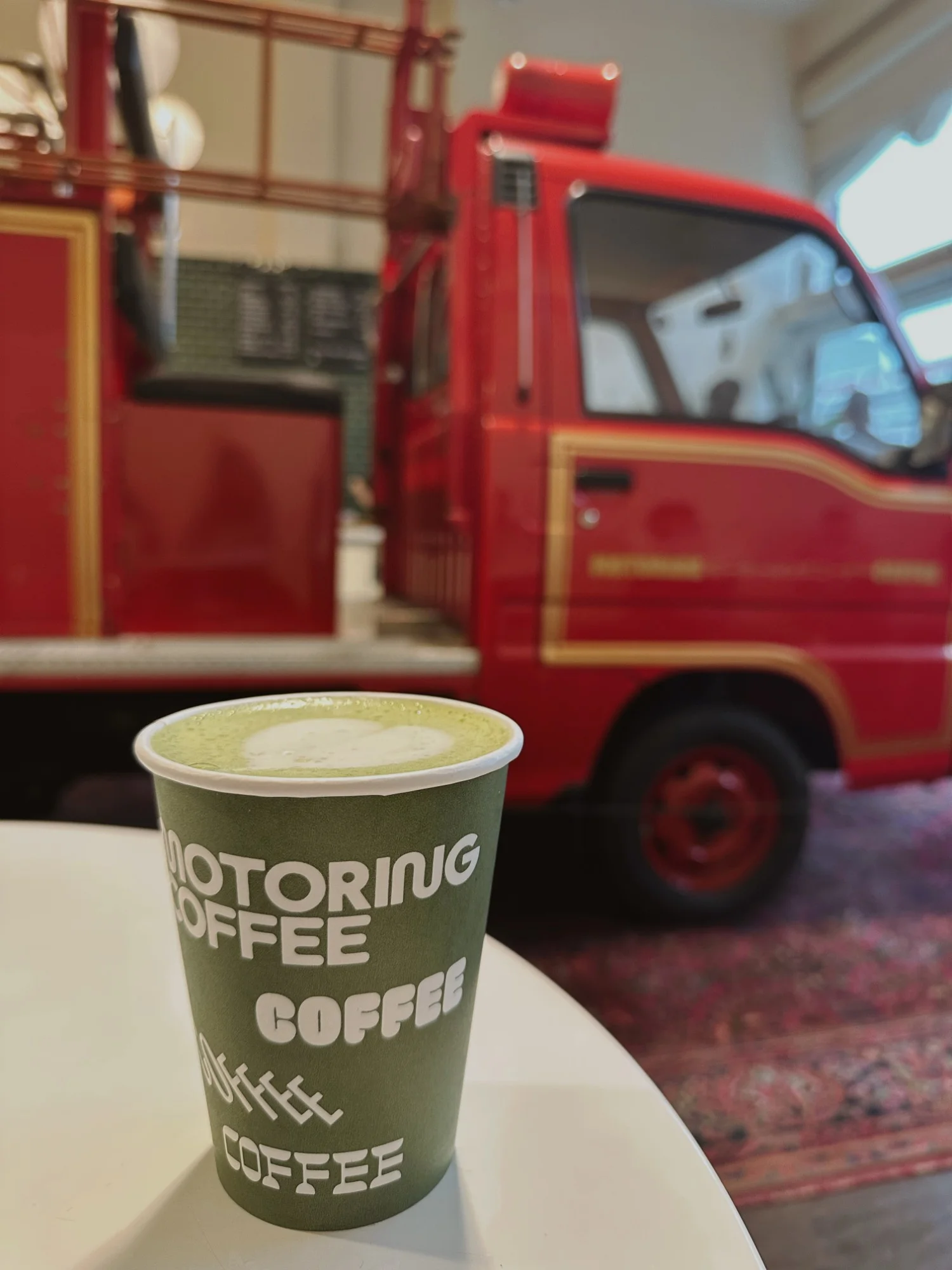 A matcha latte in a Motoring Coffee cup with a red vintage fire truck parked inside the cafe