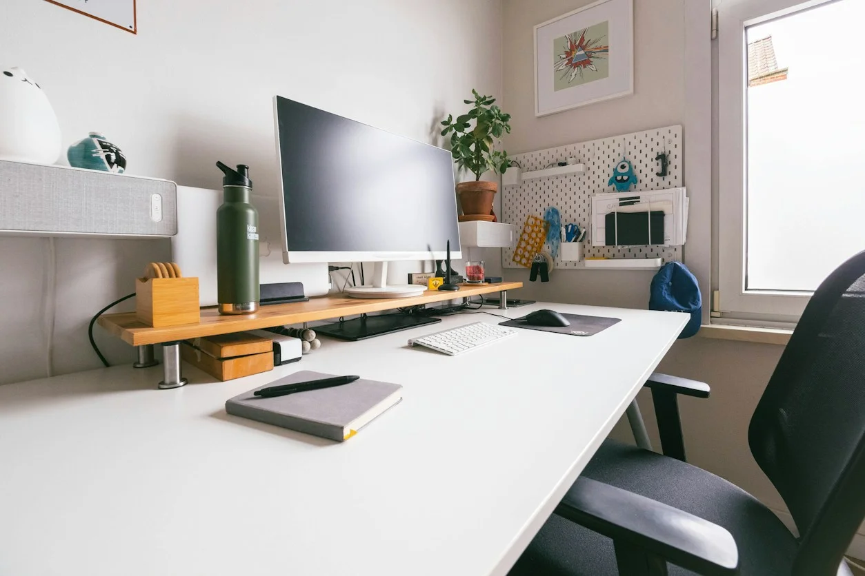 A bright white home office with a monitor on a wooden riser, a plant, and a window