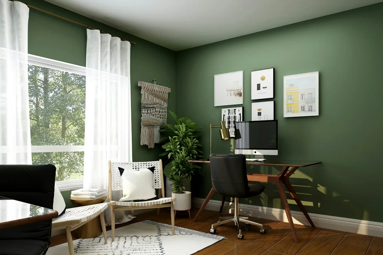 A home office with green walls, a wooden desk, a black chair, and a rattan armchair by the window