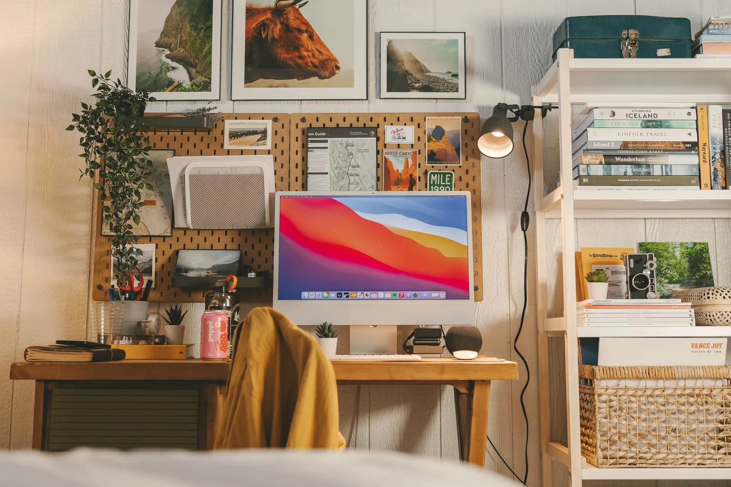 A desktop computer on a wooden desk with a pegboard of prints and maps