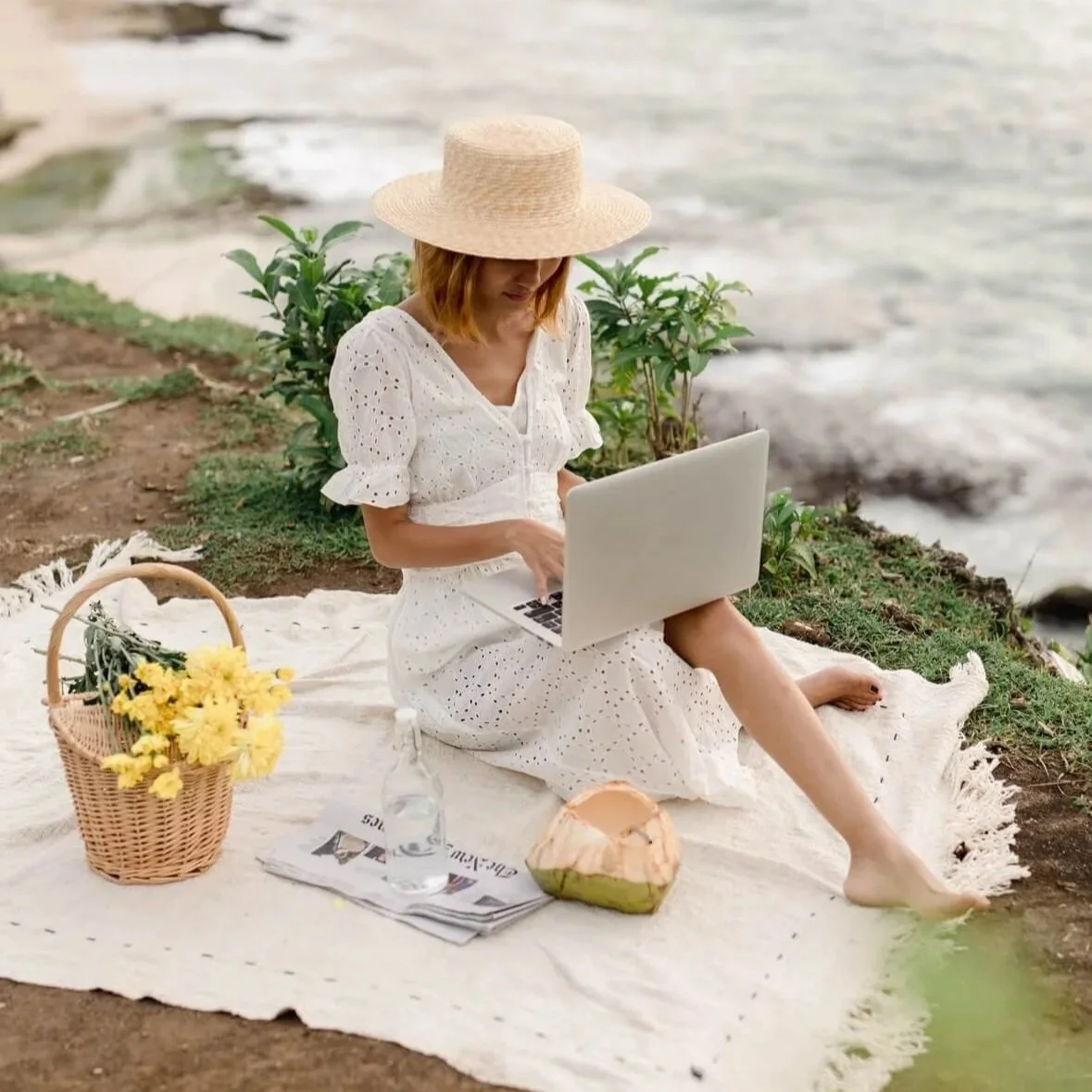 Woman in a sun hat on a picnic blanket writing on a laptop